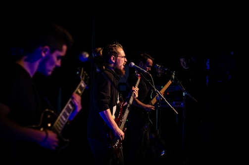 A group of musicians performing on stage, featuring a guitarist playing energetically in the foreground and a vocalist singing passionately into a microphone. The lighting is low, casting a dramatic and moody ambiance over the scene.