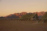Three modern, A-frame huts with slanted roofs standing in a desert landscape, with a backdrop of rugged mountains illuminated by warm, late-afternoon sunlight. The ground is sandy and arid, with sparse vegetation visible. The sky is mostly clear, casting an amber hue over the scene.