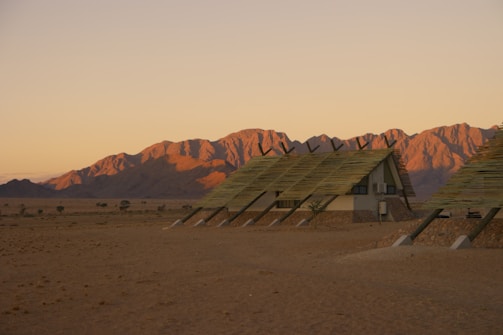 Three modern, A-frame huts with slanted roofs standing in a desert landscape, with a backdrop of rugged mountains illuminated by warm, late-afternoon sunlight. The ground is sandy and arid, with sparse vegetation visible. The sky is mostly clear, casting an amber hue over the scene.
