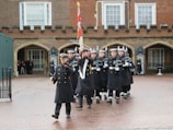 A group of uniformed individuals is marching in formation across a paved courtyard. They are dressed in dark coats and hats, some carrying rifles with bayonets. In the background, a brick building with arched doorways and large windows is visible. A few people are standing in the arches, watching the procession.