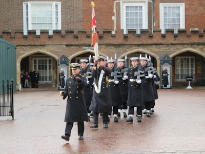 A group of uniformed individuals is marching in formation across a paved courtyard. They are dressed in dark coats and hats, some carrying rifles with bayonets. In the background, a brick building with arched doorways and large windows is visible. A few people are standing in the arches, watching the procession.