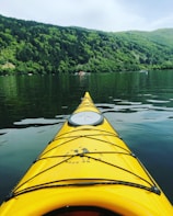 A vibrant yellow kayak is seen from the perspective of the person paddling, cutting through the calm waters of a serene lake. In the background, lush green hills covered with dense trees rise into the sky, while a small boat is visible at a distance.