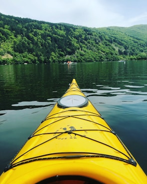 A vibrant yellow kayak is seen from the perspective of the person paddling, cutting through the calm waters of a serene lake. In the background, lush green hills covered with dense trees rise into the sky, while a small boat is visible at a distance.