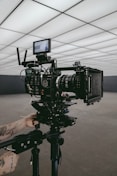 A professional video camera set up on a tripod in an indoor setting with geometric ceiling panels. A person's tattooed arms are adjusting the equipment, suggesting a behind-the-scenes look at a film production.