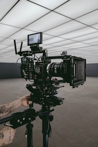 A professional video camera set up on a tripod in an indoor setting with geometric ceiling panels. A person's tattooed arms are adjusting the equipment, suggesting a behind-the-scenes look at a film production.
