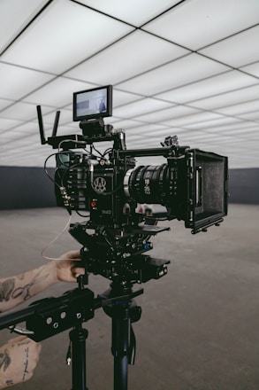 A professional video camera set up on a tripod in an indoor setting with geometric ceiling panels. A person's tattooed arms are adjusting the equipment, suggesting a behind-the-scenes look at a film production.