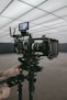 A professional video camera set up on a tripod in an indoor setting with geometric ceiling panels. A person's tattooed arms are adjusting the equipment, suggesting a behind-the-scenes look at a film production.