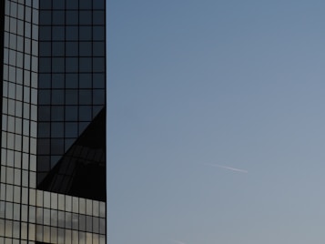 The image features a modern glass skyscraper with a reflective surface and a minimalist design. The building occupies the left side of the image and reflects the clear blue sky, with a contrail from an airplane visible in the sky on the right side.