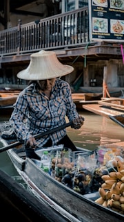 A person wearing a wide-brimmed hat navigates a wooden boat filled with various fruits, including bananas and other produce, through a bustling floating market. The boat is surrounded by water and other boats, set against a backdrop of a wooden structure with food signage.