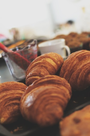 A close-up view of freshly baked, golden-brown croissants placed on a dark tray. In the background, there is a blurred image of a white coffee mug and a glass measuring cup with a red spoon. The setting suggests an inviting breakfast or brunch scene.