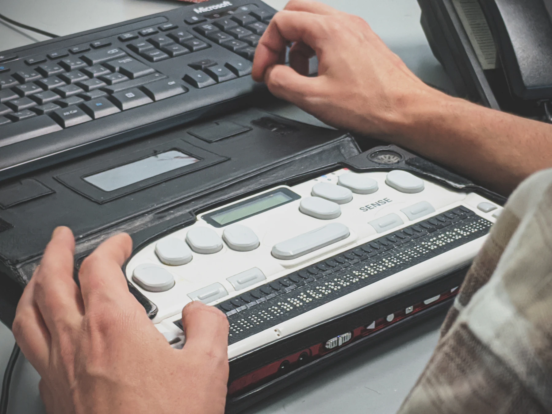 Two hands resting on a refreshable braille display, a row of pins along its front edge, with a computer keyboard behind