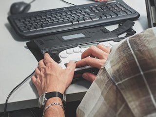 A person wearing a watch types on a Braille keyboard placed on a desk. A traditional computer keyboard and a mouse are also visible nearby. The person is wearing a plaid shirt, and the scene suggests an office or workspace setup.