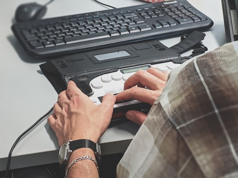 A person wearing a watch types on a Braille keyboard placed on a desk. A traditional computer keyboard and a mouse are also visible nearby. The person is wearing a plaid shirt, and the scene suggests an office or workspace setup.