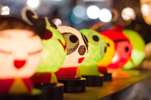 A collection of colorful, cartoonish character lamps lined up in a row. The figures feature exaggerated facial expressions and are brightly illuminated, casting a warm glow. The background is softly blurred, creating a bokeh effect with circular lights.