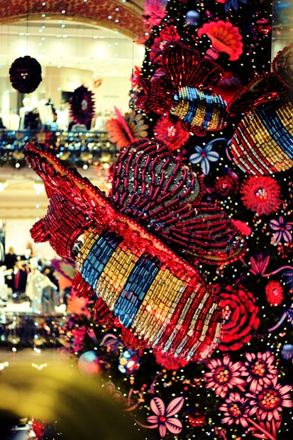 A vibrant display featuring large, colorful insect decorations with intricate patterns, surrounded by an array of vivid red and pink flowers. The background is a lively, well-lit setting with noticeable reflections and sparkling lights.