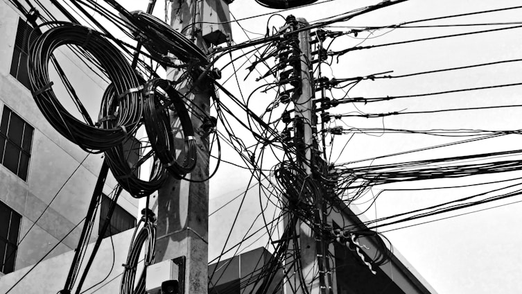 Tangled and chaotic electrical wires are attached to a utility pole in an urban setting. Large coils of cables are also visible, creating a complex network of lines against the backdrop of a building with windows.