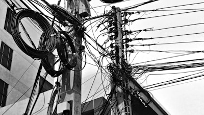 Tangled and chaotic electrical wires are attached to a utility pole in an urban setting. Large coils of cables are also visible, creating a complex network of lines against the backdrop of a building with windows.