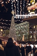 A festive nighttime scene at a holiday market, featuring a tall Christmas tree adorned with numerous lights. Strings of twinkling lights hang overhead, creating a warm and cheerful atmosphere. People bundled in winter clothing walk among stalls with striped canopies, enjoying the festive ambiance. A modern building with glass windows is visible in the background.