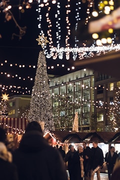 A festive nighttime scene at a holiday market, featuring a tall Christmas tree adorned with numerous lights. Strings of twinkling lights hang overhead, creating a warm and cheerful atmosphere. People bundled in winter clothing walk among stalls with striped canopies, enjoying the festive ambiance. A modern building with glass windows is visible in the background.