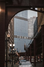A city scene features a large building with glass exteriors, prominently displaying the word 'TRUMP' across its facade. The foreground includes a busy street with cars and pedestrians. Overhead train tracks and lampposts frame the street, contributing to the bustling urban atmosphere.