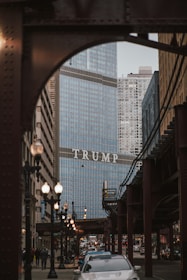 A city scene features a large building with glass exteriors, prominently displaying the word 'TRUMP' across its facade. The foreground includes a busy street with cars and pedestrians. Overhead train tracks and lampposts frame the street, contributing to the bustling urban atmosphere.