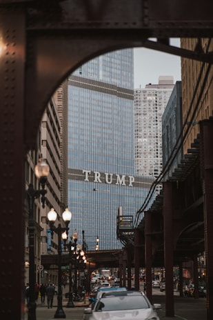 A city scene features a large building with glass exteriors, prominently displaying the word 'TRUMP' across its facade. The foreground includes a busy street with cars and pedestrians. Overhead train tracks and lampposts frame the street, contributing to the bustling urban atmosphere.