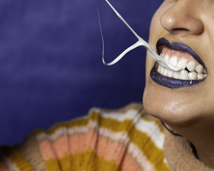 A close-up of a person with dark blue lipstick, stretching a piece of gum between their teeth. The person is wearing a multicolored striped sweater, and the background is a deep blue.