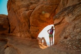 A person in outdoor gear stands on a rock formation, overlooking a natural arch. The scene is set in a desert landscape with reddish-brown rock formations.