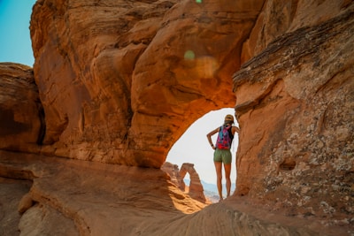 A person in outdoor gear stands on a rock formation, overlooking a natural arch. The scene is set in a desert landscape with reddish-brown rock formations.