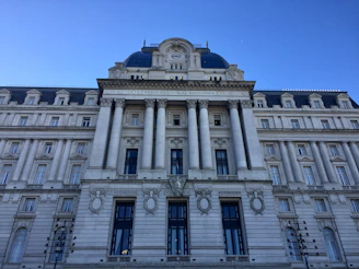 A grand neoclassical building features ornate architecture with large columns, decorative stonework, and numerous windows. The facade displays a sign indicating it is the 'Centro Cultural Kirchner'. The sky is clear, providing a vivid blue backdrop.