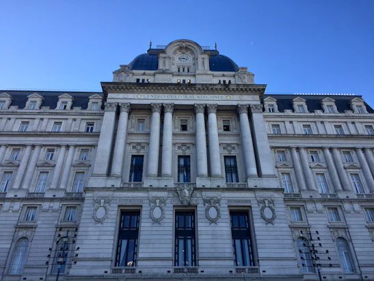 A grand neoclassical building features ornate architecture with large columns, decorative stonework, and numerous windows. The facade displays a sign indicating it is the 'Centro Cultural Kirchner'. The sky is clear, providing a vivid blue backdrop.