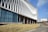 A modern architectural building with an intricate facade featuring vertical white slats. Large glass windows allow natural light in, and there is a person walking on a path surrounded by dry grass. The sky is clear and blue, suggesting a sunny day.