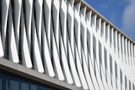 A modern architectural facade featuring a series of vertical, angled white panels evenly spaced along the building's exterior. The panels create an interplay of light and shadows, adding a dynamic and sculptural quality to the structure. The blue sky is visible in the background, enhancing the design's contrast.