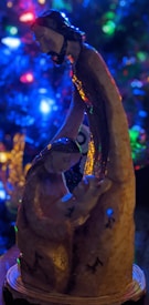 A nativity scene figurine featuring three figures is depicted. The figures are artistically styled with a focus on two adults and a child, likely representing the Holy Family. The background is a blur of colorful lights, primarily blue, red, and green, possibly from a decorated Christmas tree.