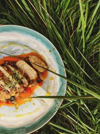 A plate of sliced grilled chicken with a garnish of microgreens rests on a bed of vibrant orange puree and assorted vegetables. The setting includes lush green grass surrounding the dish, providing a natural and earthy background.