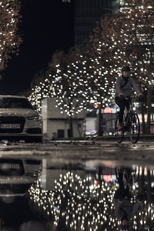 A nighttime urban scene with bright, festive lights adorning the trees, creating a warm glow. A person is riding a bicycle on a wet street, which reflects the lights and the cyclist, adding depth and symmetry. A parked car is visible on the side of the road.