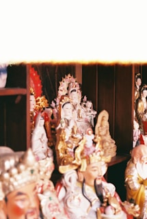 A collection of ornate and colorful figurines is displayed on shelves. These figures appear to be intricately crafted and depict various deities or spiritual icons, showcasing detailed costumes and diverse expressions. The background consists of a wooden cabinet, which adds a warm tone to the setting.