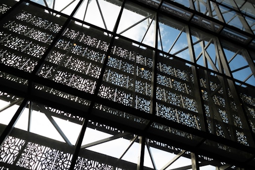 Intricate metallic lattice patterns framed within a transparent glass structure, allowing glimpses of the blue sky through geometric designs.