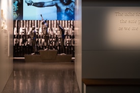 A dimly lit display features statues of athletes standing on a podium. They are positioned in front of a backdrop with the words 'LEVELING THE PLAYING FIELD' in bold letters. On the wall to the right, a softly lit quote is partially visible. The overall ambiance suggests a museum or exhibition setting.