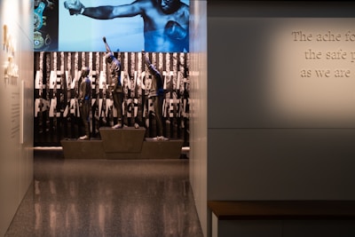 A dimly lit display features statues of athletes standing on a podium. They are positioned in front of a backdrop with the words 'LEVELING THE PLAYING FIELD' in bold letters. On the wall to the right, a softly lit quote is partially visible. The overall ambiance suggests a museum or exhibition setting.