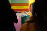 A vibrant and colorful exhibit with a circular arrangement of blocks or panels in various colors. Two people are partially visible in the foreground. The wall features horizontal stripes of red, green, yellow, and orange, with text that reads 'Pick a Color, Any Flavor.'