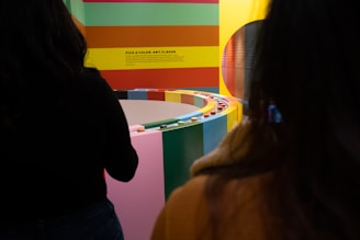 A vibrant and colorful exhibit with a circular arrangement of blocks or panels in various colors. Two people are partially visible in the foreground. The wall features horizontal stripes of red, green, yellow, and orange, with text that reads 'Pick a Color, Any Flavor.'
