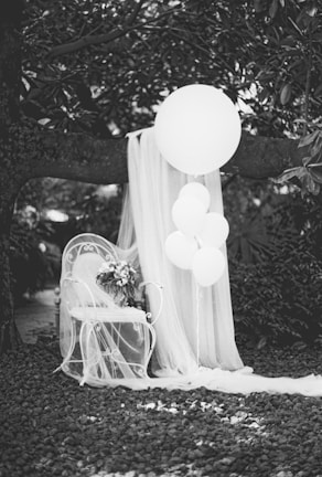 An elegant outdoor scene with a white wrought iron chair decorated with a flowing sheer fabric and a bouquet of flowers. A cluster of white balloons is tied to the chair, while a larger balloon and more fabric hang from a tree branch. The ground is covered with small pebbles and surrounding foliage adds a natural backdrop.