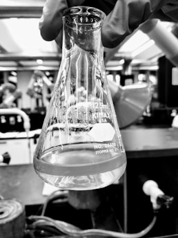 A gloved hand holding a 125ml conical flask with a label indicating it is from Kimax. The flask contains a transparent liquid with some residue at the bottom. The background appears to be a laboratory setting with various scientific equipment and blurred figures.