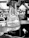 A gloved hand holding a 125ml conical flask with a label indicating it is from Kimax. The flask contains a transparent liquid with some residue at the bottom. The background appears to be a laboratory setting with various scientific equipment and blurred figures.