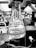 A gloved hand holding a 125ml conical flask with a label indicating it is from Kimax. The flask contains a transparent liquid with some residue at the bottom. The background appears to be a laboratory setting with various scientific equipment and blurred figures.
