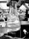 A gloved hand holding a 125ml conical flask with a label indicating it is from Kimax. The flask contains a transparent liquid with some residue at the bottom. The background appears to be a laboratory setting with various scientific equipment and blurred figures.