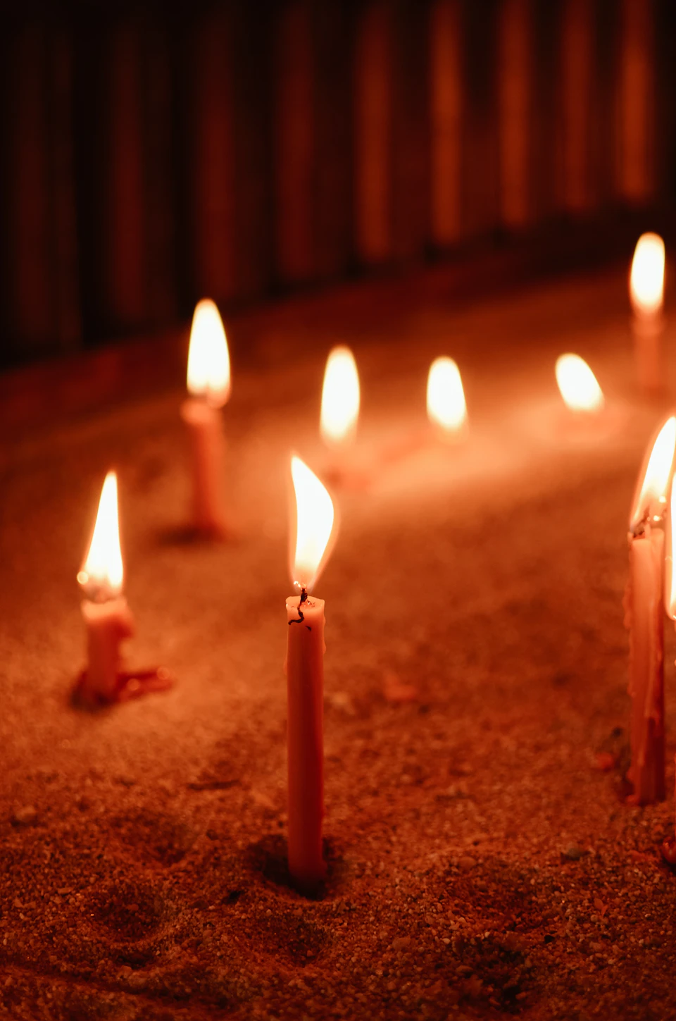 The image shows a group of lit candles arranged on a sandy surface, with their flames dancing and casting a warm glow. The candles are positioned at various angles, creating an interesting visual effect. In total, there are eight candles visible in the scene, each one burning brightly against the dark background.