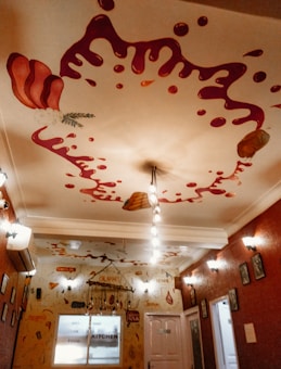 A creatively decorated hallway features walls adorned with drawings of food items and sauces. The ceiling is painted with splashes of red sauce and bread, giving a whimsical and artistic vibe. Light fixtures hang from the ceiling, and the walls have framed artwork and various culinary words and phrases written on them.