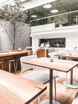 A modern, minimalist cafe interior with light wood tables and white mesh chairs. A tree is positioned indoors near the center, and the space features a mezzanine level with glass railings. Two people are visible in the open kitchen area wearing uniforms, with kitchen utensils hanging on the wall. The lighting is soft and natural, coming through large windows or skylights.